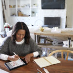 Elevated view of middle aged mixed race woman sitting at a table in her dining room using a stylus with a tablet computer, elevated view, focus on foreground